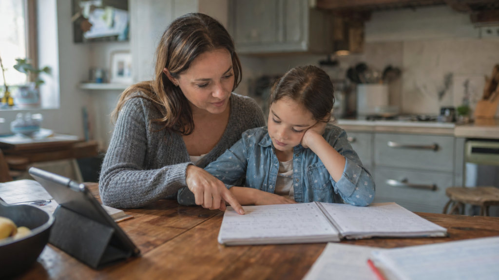 Parent sitting beside a child at a kitchen table, gently guiding homework with questions while the child works in a notebook.